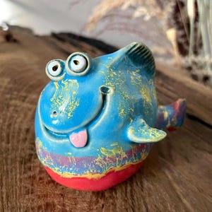May include: A whimsical ceramic fish figurine in shades of blue, yellow, red, and purple. The fish has large, googly eyes, a smiling mouth with a pink tongue, and small fins. The figurine is hand-painted with a textured finish.
