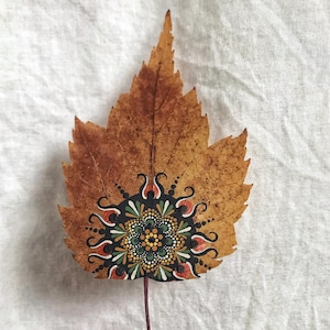 May include: A dried, brown leaf with a detailed, hand-painted mandala design in black, red, green, and yellow. The mandala is centered on the leaf, showcasing intricate patterns. The leaf is held by a person with a blue painted fingernail.