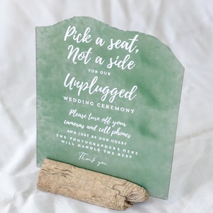 May include: A wedding sign with a green watercolour background and white text. The sign reads "Pick a seat, Not a side for our Unplugged Wedding Ceremony." It is supported by a piece of driftwood.