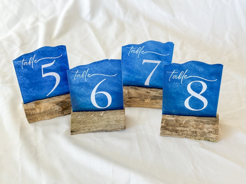 Handcrafted acrylic table numbers spray painted to mimic the appearance of sea glass, mounted on rustic driftwood stands. Perfect for beach-themed weddings and coastal events, these unique table numbers blend natural elements with artistic design.