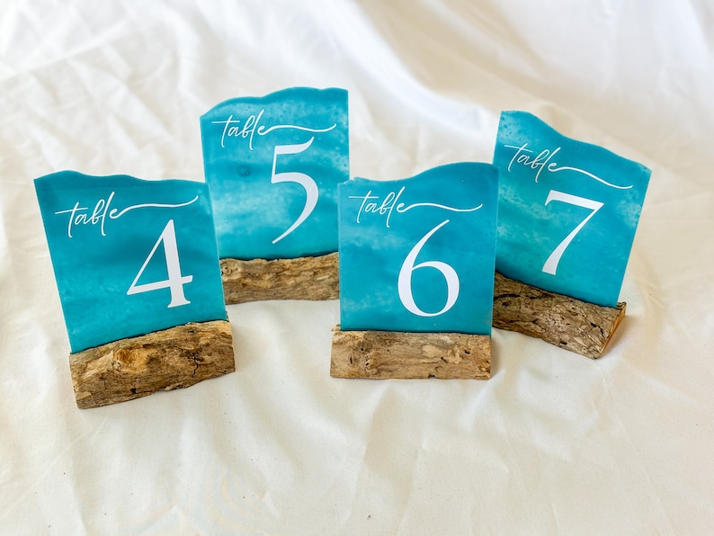 Handcrafted acrylic table numbers spray painted to mimic the appearance of sea glass, mounted on rustic driftwood stands. Perfect for beach-themed weddings and coastal events, these unique table numbers blend natural elements with artistic design.