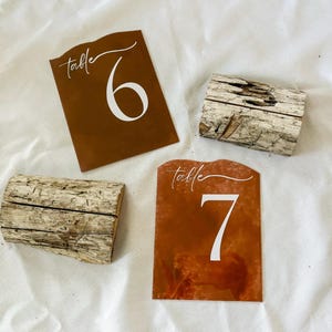 May include: Two rustic wooden table number holders with brown and white table number cards. The cards have the text "table" and the numbers "6" and "7" in white.