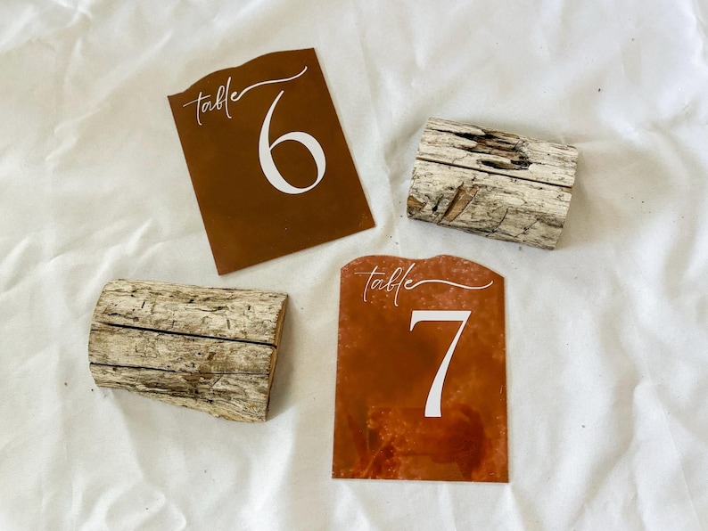 May include: Two rustic wooden table number holders with brown and white table number cards. The cards have the text "table" and the numbers "6" and "7" in white.