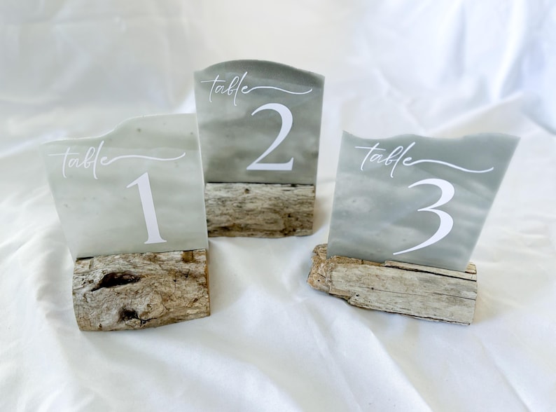 May include: Three table number signs with the words "table" and the numbers 1, 2, and 3 written in white on a light gray background. The signs are on top of small pieces of driftwood.