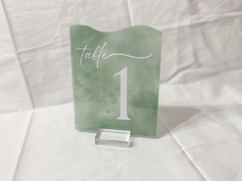 May include: Green table number card with a white acrylic stand. The card has a white cursive font that says "Table" and a white number "1" on a green background.