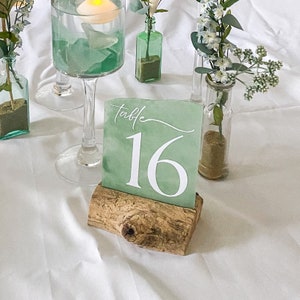 Handcrafted acrylic table numbers spray painted to mimic the appearance of sea glass, mounted on rustic driftwood stands. Perfect for beach-themed weddings and coastal events, these unique table numbers blend natural elements with artistic design.