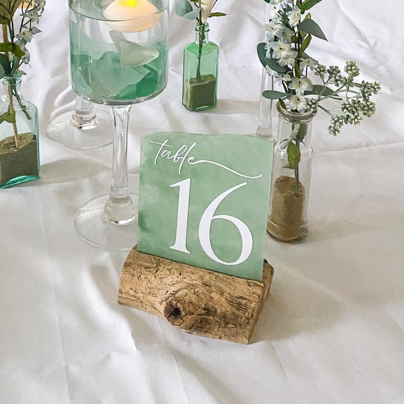 Handcrafted acrylic table numbers spray painted to mimic the appearance of sea glass, mounted on rustic driftwood stands. Perfect for beach-themed weddings and coastal events, these unique table numbers blend natural elements with artistic design.
