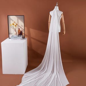 May include: A white bridal cape with a high neckline and a long, flowing train. The cape is draped over a mannequin, showcasing its elegant design. The fabric appears to be lightweight and sheer, creating a graceful silhouette.