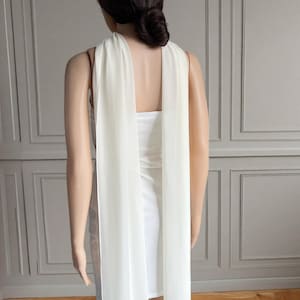 Op de afbeelding: Een witte strapless minijurk met twee lange, vloeiende stoffen panelen bevestigd aan de achterkant van de halslijn die tot aan de zoom vallen. Het kledingstuk is gemaakt van een gladde, ondoorzichtige stof met doorschijnende panelen.