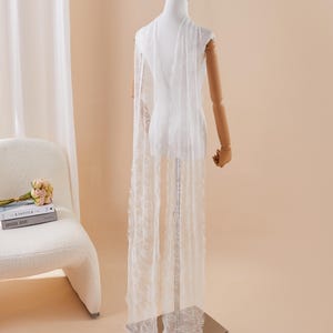 Puede incluir: Una capa blanca y transparente con bordes deshilachados está drapeada sobre un maniquí. La capa sin mangas llega hasta el suelo. El maniquí tiene brazos de madera y está sobre un soporte metálico. El fondo es de color neutro.