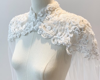 Puede incluir: Una capa de novia de encaje blanco con cuello alto y mangas largas. La capa está adornada con lentejuelas y cuentas.