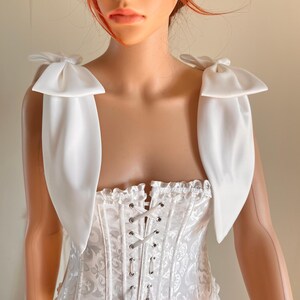 Può includere: Corsetto bianco in satin con lacciatura sul davanti e un fiocco di raso bianco su ogni spallina.