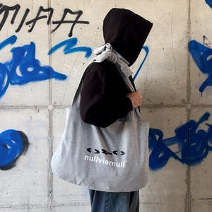 May include: A large grey tote bag made of soft fabric, featuring a knotted shoulder strap and the black printed text "nullviernull" with a stylized graphic symbol above it. The bag is worn over the shoulder, displaying a simple, functional design.