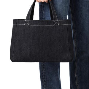 Può includere: Una borsa tote in denim nero con dettagli di cuciture bianche. La borsa ha due manici e una forma rettangolare. Le cuciture sono visibili lungo la parte superiore e sugli attacchi dei manici. La borsa è tenuta da una persona che indossa jeans blu.