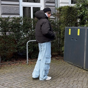 Puede incluir: Una persona lleva una chaqueta acolchada negra con capucha, un gorro negro y gafas de sol. Lleva unos vaqueros azules claros con bolsillos cargo y zapatillas blancas.