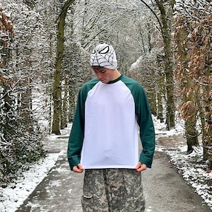 May include: A white long-sleeve baseball tee with green sleeves. The person is wearing a white beanie with a black spiderweb design.
