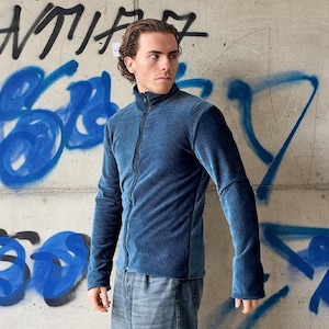 Könnte beinhalten: Eine blaue Cordjacke mit Reißverschluss, hohem Kragen und langen Ärmeln. Das Kleidungsstück ist durchgehend mit einer vertikalen Rippenstruktur an Körper und Ärmeln versehen und wird mit einem zentralen Reißverschluss vom Saum bis zum Hals geschlossen.
