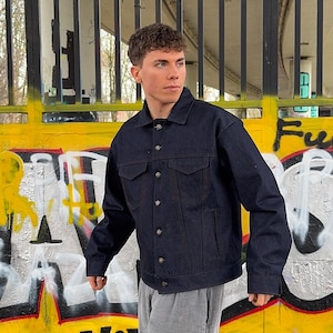 Op de afbeelding: Een donkerblauwe spijkerjas met een klassiek ontwerp. De jas heeft twee borstzakken, twee zijzakken en knoopsluitingen. De stiksels zijn in een contrasterende oranje kleur. De jas wordt door een persoon gedragen.