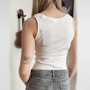 May include: A white ribbed tank top. The sleeveless top has a scoop neck and a fitted silhouette. The model is wearing gray denim jeans. A small tattoo is visible on the left arm. A ukulele hangs on the wall in the background.