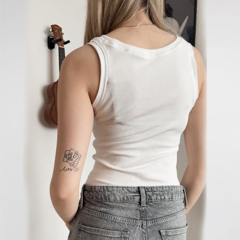 May include: A white ribbed tank top. The sleeveless top has a scoop neck and a fitted silhouette. The model is wearing gray denim jeans. A small tattoo is visible on the left arm. A ukulele hangs on the wall in the background.