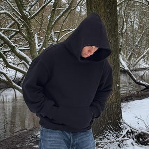 Op de afbeelding: Een zwarte hoodie met een kangoeroezak. De persoon draagt een blauwe spijkerbroek. De achtergrond toont een besneeuwd bos met een rivier en kale bomen.