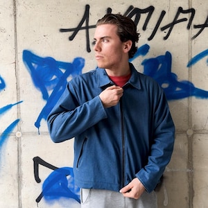 May include: A blue zip-up jacket with a classic collar and long sleeves, worn over a red shirt. The jacket features a central black zipper closure and two side pockets. The fabric has a solid blue tone with a subtle texture, providing a clean look.