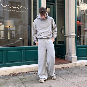 Op de afbeelding: Een grijs hoodie met een kangoeroezak en een bijpassende grijze joggingbroek. De persoon staat voor een winkel met een bord waarop "Lampenschirme und Leuchten" staat.