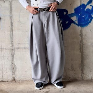 Op de afbeelding: Lichtgrijze broek met wijde pijpen, diepe plooien aan de voorzijde, een hoge taille en een zwarte riem met een zilverkleurige gesp. De broek wordt gecombineerd met een wit shirt met lange mouwen en zwarte sneakers met witte zolen, volledig beeld.