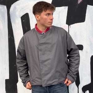Puede incluir: Chaqueta gris con cierre cruzado. La chaqueta tiene un cuello rojo y se usa sobre una camisa estampada. El fondo es una pared de graffiti en blanco y negro.