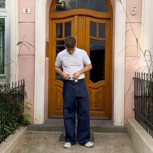 Puede incluir: Una persona vestida con una camiseta blanca con un logotipo y unos vaqueros anchos de color azul oscuro. La persona está de pie frente a una puerta de madera y un edificio rosa.