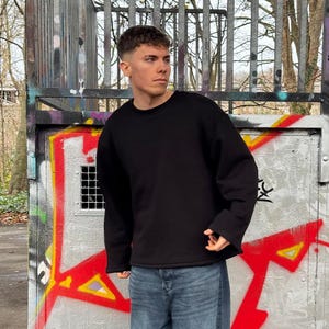 Könnte beinhalten: Ein einfarbiges, schwarzes Langarm-Sweatshirt mit Rundhalsausschnitt und breiten Bündchen, kombiniert mit einer blauen Jeans. Das Sweatshirt hat ein schlichtes, musterloses Design mit lockerer Passform, während die Jeans eine klassische Waschung aufweist.