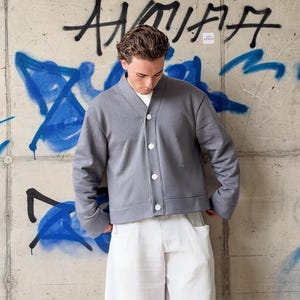May include: A grey long-sleeve cardigan with a V-neckline and a front button closure featuring four white buttons. The garment has a structured collar, wide cuffs, and a straight hemline, worn over a plain white shirt and white trousers.