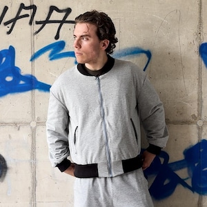 Könnte beinhalten: Eine hellgraue Bomberjacke mit schwarzem Rippstrickkragen, schwarzen Rippstrickbündchen und einem schwarzen Rippstricksaum. Die Jacke hat einen silbernen Reißverschluss in der Mitte und zwei Seitentaschen. Der Stoff wirkt wie weicher Jersey.