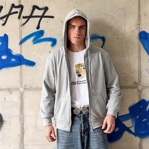 Puede incluir: Una sudadera con capucha y cremallera en color gris claro, con cordones y dos bolsillos delanteros, sobre una camiseta blanca con un gráfico de escudo dorado y negro con el texto 200, combinada con vaqueros azules y un cinturón negro.