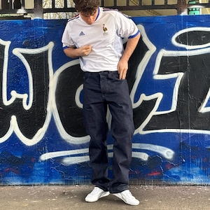 May include: A person wearing a white soccer jersey with blue accents and dark blue jeans. The jersey has a logo on the chest. The person is wearing white sneakers. The background is a graffiti-covered wall.
