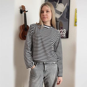 Op de afbeelding: Een shirt met lange mouwen en een horizontaal zwart-wit gestreept patroon met een ronde hals. Het shirt wordt gecombineerd met een grijze spijkerbroek met een klassiek ontwerp met vijf zakken en een knoopsluiting voor een casual outfit.