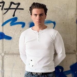 May include: A white long-sleeved henley shirt featuring a ribbed texture, a three-button placket at the neckline, and a circular white patch with three black dots positioned on the lower right side of the torso.