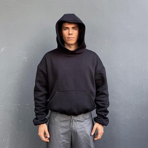 May include: A black hooded sweatshirt with a kangaroo pocket. The sweatshirt is worn by a person in front of a gray wall. The sleeves are cuffed at the wrists. The person is wearing gray pants.