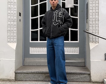 May include: A black zip-up jacket featuring a white geometric line pattern on the chest, paired with loose-fitting blue denim jeans and light-coloured athletic trainers. The jacket has a hood and long sleeves, while the jeans have a wide-leg cut.