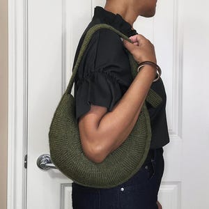 May include: Olive green knitted crescent-shaped shoulder bag. The bag has a ribbed texture and a long strap. The person in the image is wearing a black top and dark blue jeans. The bag is carried over the shoulder.