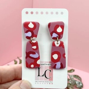 May include: A pair of handmade, abstract polymer clay earrings. The earrings are a mauve color with red, white, and purple abstract shapes. The earrings are attached to a white card with the text "yatively LC clay creatively handcrafted".