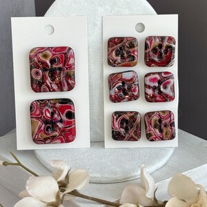 Puede incluir: Botones cuadrados con diseño veteado en rojo, negro, dorado y blanco. Los botones están sujetos a tarjetas blancas. Los botones tienen cuatro agujeros para coser. Suministros de costura.