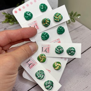 May include: A selection of green and gold knot-shaped stud earrings on white cards. Some earrings feature silver detailing. The cards are printed with "clayatively" and "LC". The earrings are presented against a blurred backdrop.