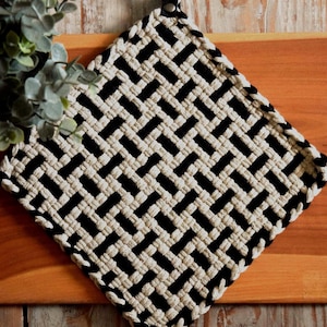 May include: A square potholder featuring a woven basket-weave pattern in alternating black and cream-coloured fabric strips. The potholder has a small black loop at the top corner for hanging and is constructed from thick, textured textile material.