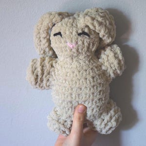 Puede incluir: Un peluche de conejo beige tejido a ganchillo con orejas caídas, ojos cosidos negros y una nariz rosa. El peluche hecho a mano está sujeto por una mano y tiene un aspecto suave y texturizado. El conejo es un juguete lindo y tierno.