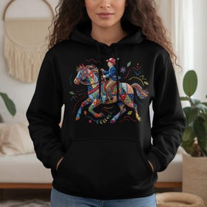 Op de afbeelding: Zwarte hoodie met een kleurrijke afbeelding van een paard en ruiter. Het paard heeft een patchwork-ontwerp in diverse kleuren, en de ruiter draagt een blauwe jas en rode hoed. Het ontwerp bevat bloemen- en steraccenten.