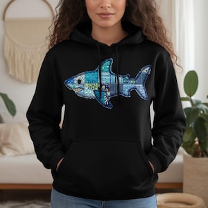 May include: Black hooded sweatshirt featuring a detailed, patchwork-style shark design in shades of blue, teal, and white. The shark graphic is centered on the front of the hoodie. The sweatshirt has a kangaroo pocket and a drawstring hood.