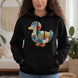Könnte beinhalten: Schwarzer Kapuzenpullover mit einem bunten Patchwork-Enten-Design. Die Ente hat einen gelben Schnabel, orangefarbene Füße und einen Körper aus verschiedenen Stoffmustern in Rot, Blau, Grün und Gelb. Der Pullover hat eine Kängurutasche.