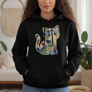 Op de afbeelding: Zwarte hoodie met een kleurrijk patchwork kattenmotief. De kat is gemaakt van verschillende stoffen met patronen in blauw, rood en geel. De sweatshirt heeft een kangoeroezak en een capuchon met trekkoord.