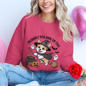 Może przedstawiać: Różowa bluza z napisem "PROBABLY DOG HAIR ON THIS". Grafika przedstawia kreskówkowego corgi w kapeluszu i pelerynie czarownicy, jadącego na miotle, z dyniami, duchami i nietoperzami.